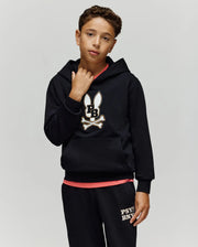 A boy wears the Psycho Bunny KIDS WADE CORD PATCH HOODIE (KT0500031), a black premium cotton blend hoodie with an embroidered Bunny patch, and black sweatpants, standing against a plain background, looking at the camera with one hand on his collar.