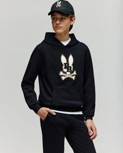 A young person wears the Psycho Bunny KIDS WADE CORD PATCH HOODIE - KT0500031 in black with an embroidered Bunny and crossbones logo, paired with black pants and cap, standing against a plain light background with one hand in their pocket.