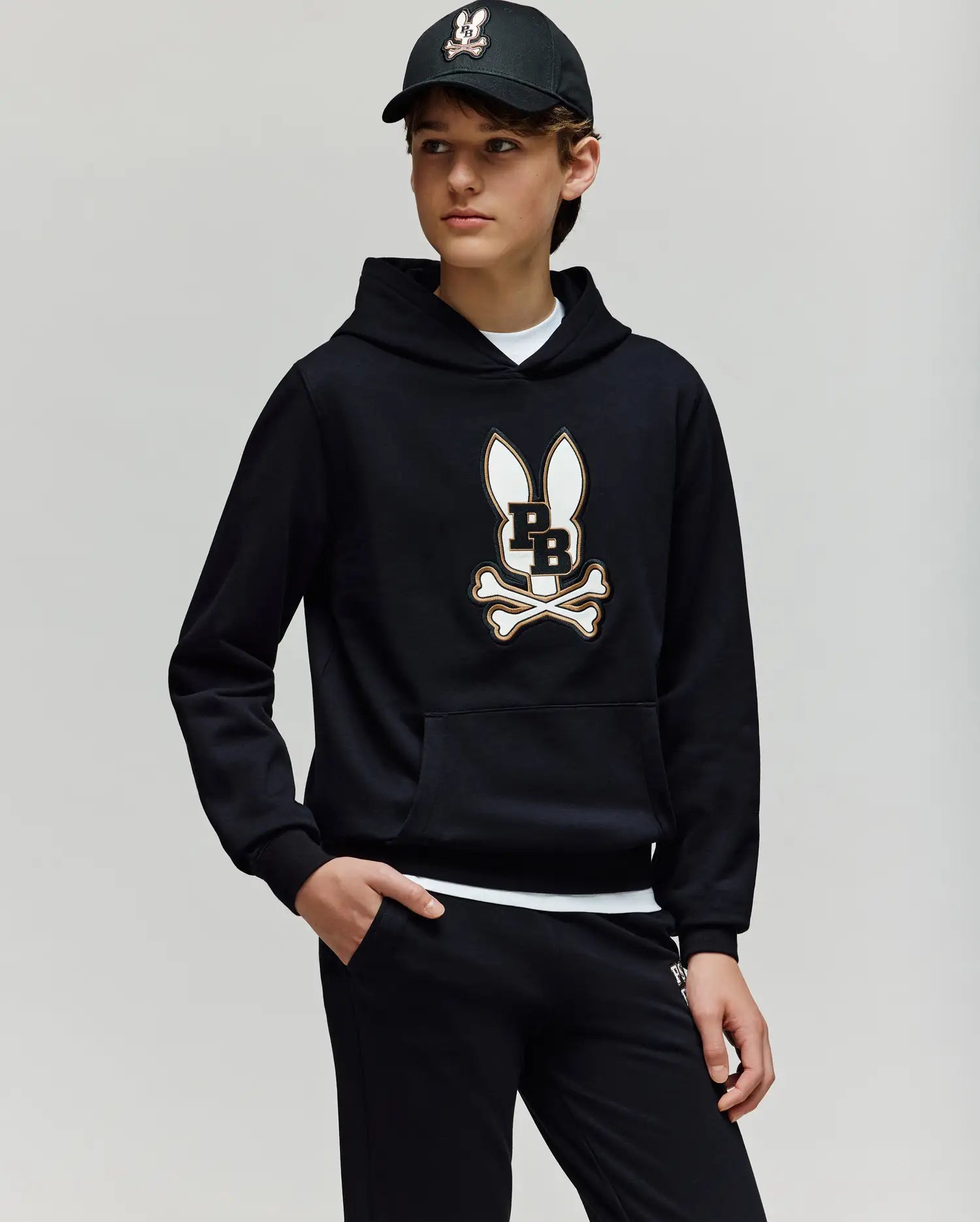 A young person wears the Psycho Bunny KIDS WADE CORD PATCH HOODIE - KT0500031 in black with an embroidered Bunny and crossbones logo, paired with black pants and cap, standing against a plain light background with one hand in their pocket.