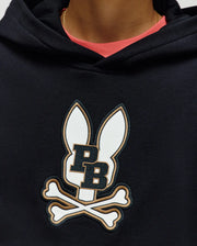 A person wears the Psycho Bunny KIDS WADE CORD PATCH HOODIE (KT0500031), made from a premium cotton blend with an embroidered Bunny patch—two ears, "PB," and crossed bones. The hood is up, showing a coral shirt and gold necklace underneath.