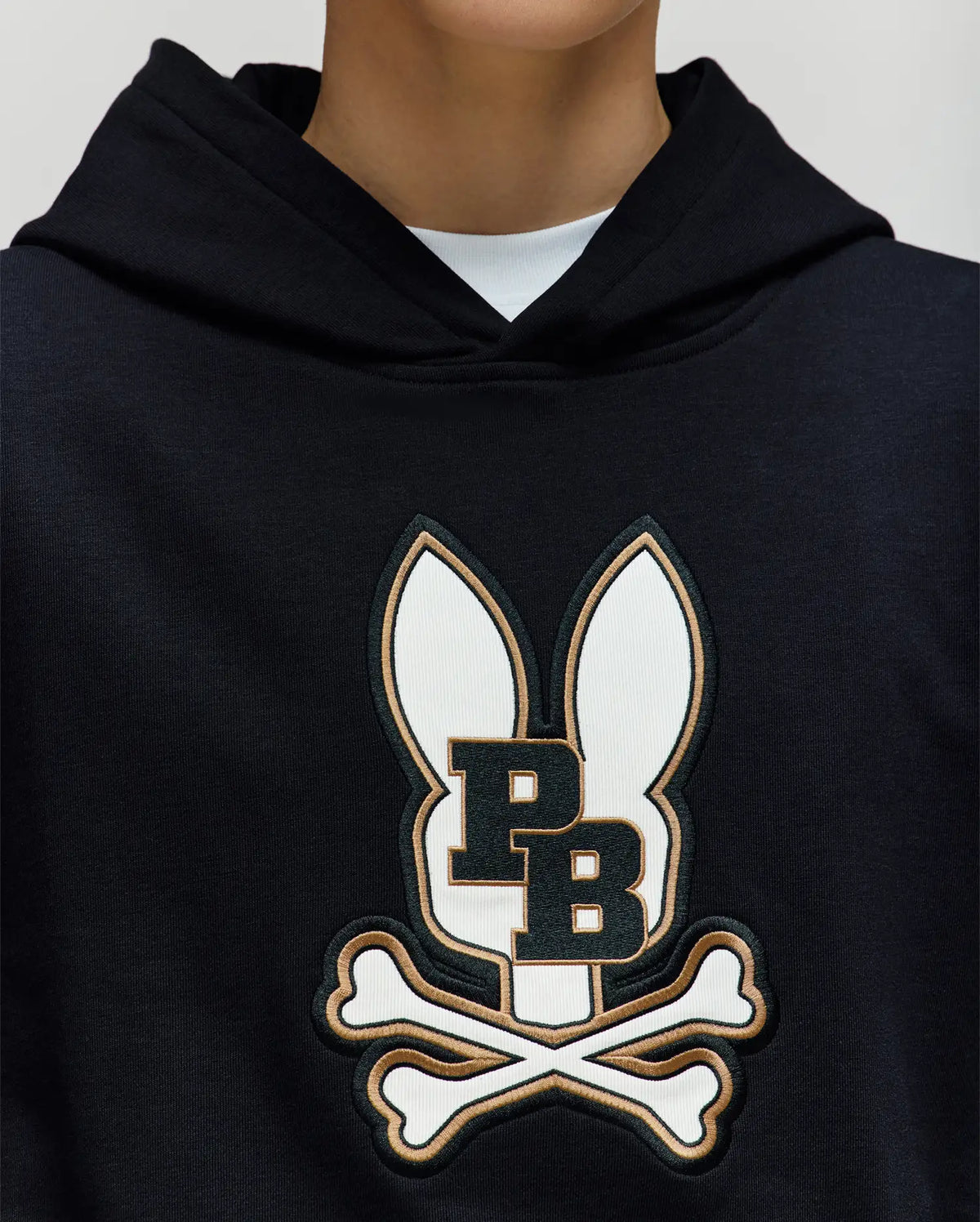A person wears the Psycho Bunny KIDS WADE CORD PATCH HOODIE (KT0500031), crafted from a premium cotton blend and featuring a large embroidered Bunny patch with ears, "P B" lettering, and crossed bones; only their lower face and neck are visible.
