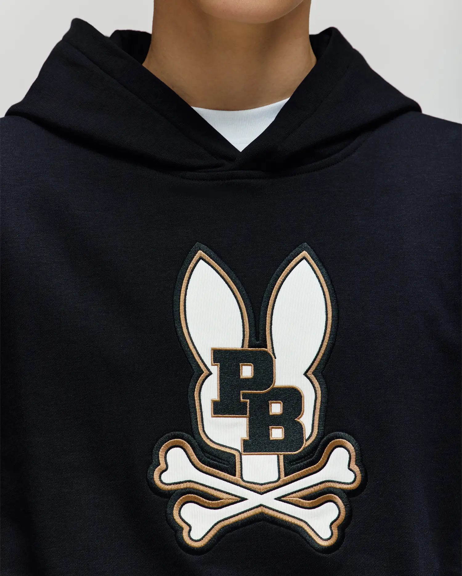 A person wears the Psycho Bunny KIDS WADE CORD PATCH HOODIE (KT0500031), crafted from a premium cotton blend and featuring a large embroidered Bunny patch with ears, "P B" lettering, and crossed bones; only their lower face and neck are visible.