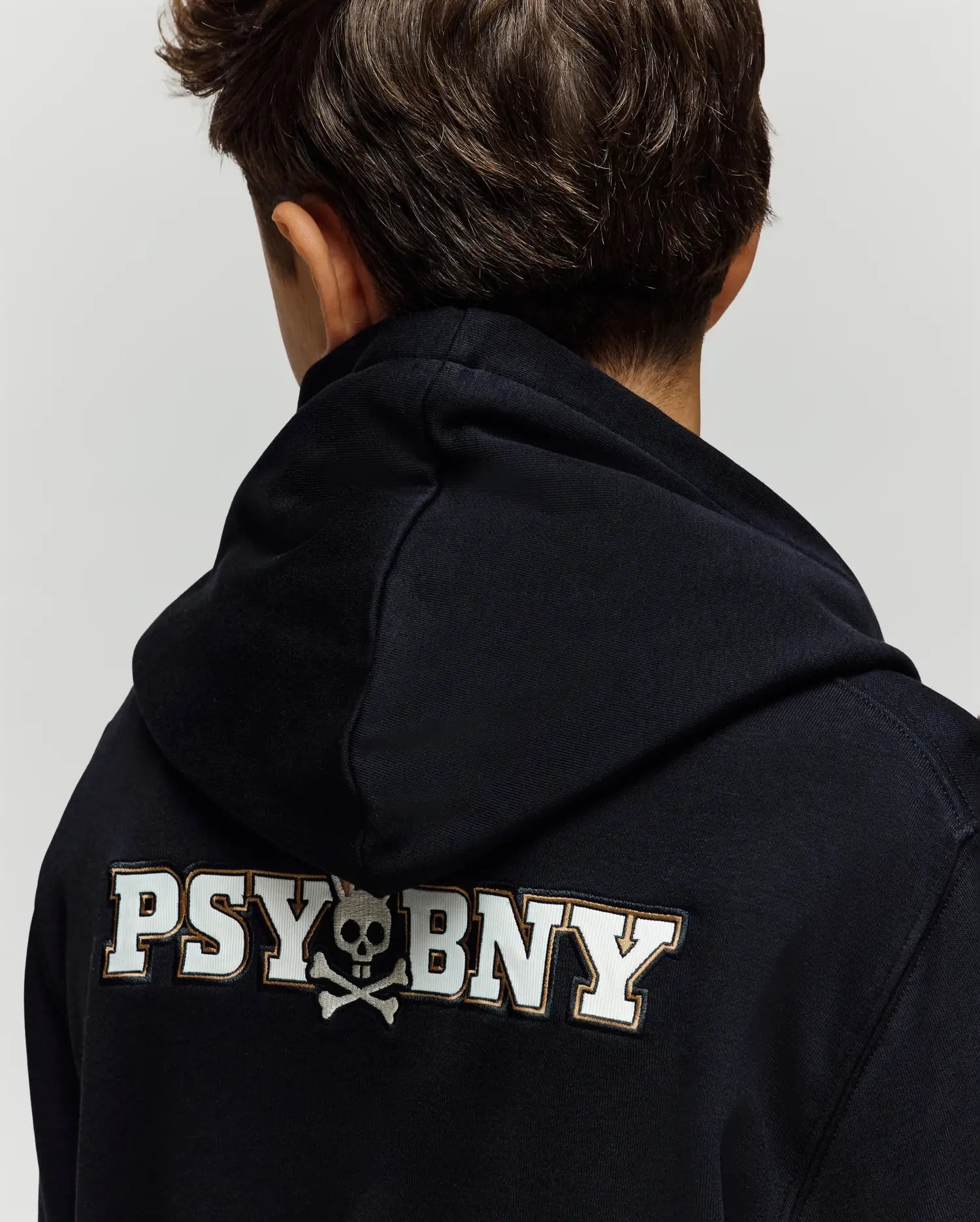 A child with short brown hair is seen from behind wearing the Psycho Bunny KIDS WADE CORD PATCH HOODIE, featuring "PSYBNY" and a skull and crossbones graphic on the back, crafted from a premium cotton blend for comfort.