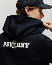A child wearing the Psycho Bunny KIDS WADE CORD PATCH HOODIE (KT0500031) faces away, lifting the hoodie to reveal a "PSYBNY" graphic with a bunny-eared skull and crossbones, made from a premium cotton blend for comfort.