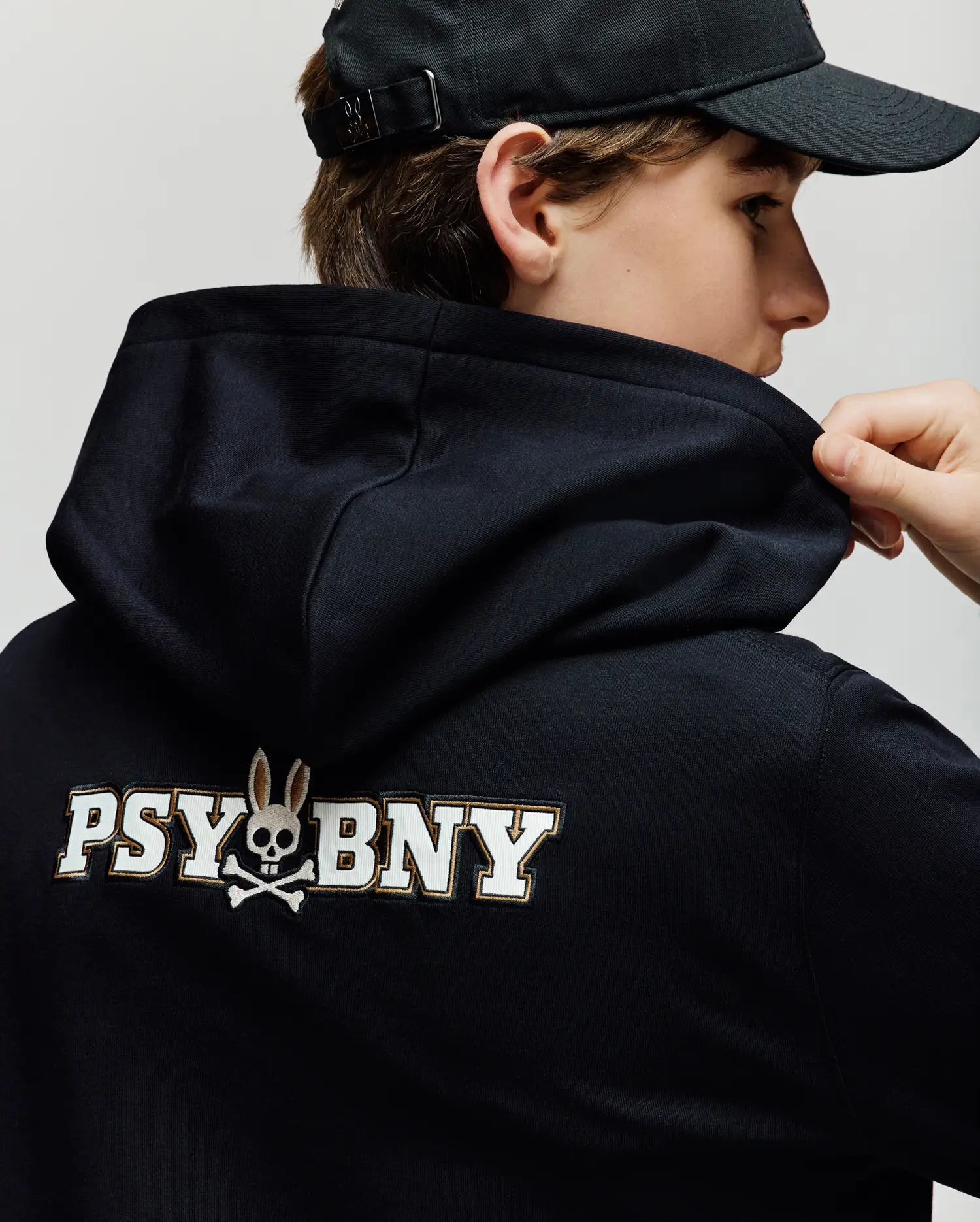 A child wearing the Psycho Bunny KIDS WADE CORD PATCH HOODIE (KT0500031) faces away, lifting the hoodie to reveal a "PSYBNY" graphic with a bunny-eared skull and crossbones, made from a premium cotton blend for comfort.