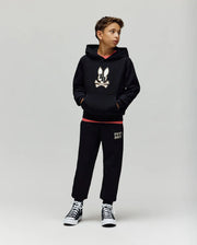 A young boy stands against a plain background, wearing the Psycho Bunny KIDS WADE CORD PATCH HOODIE (KT0500031), black pants, and black high-top sneakers with white soles, his hands tucked in his hoodie pockets.