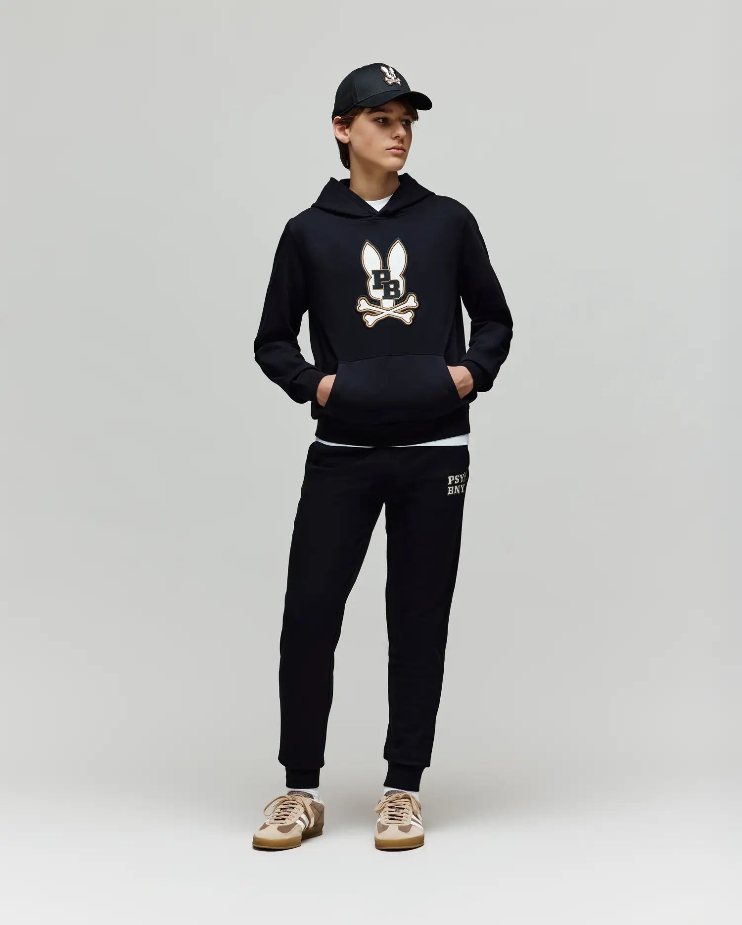 A child models the Psycho Bunny KIDS WADE CORD PATCH HOODIE (KT0500031) paired with black joggers, tan and white sneakers, and a black cap, standing against a plain light gray background.