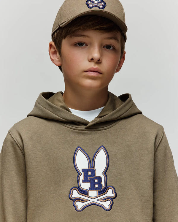 A young boy wears the Psycho Bunny KIDS WADE CORD PATCH HOODIE (KT0500031), a tan cotton blend athletic hoodie featuring an embroidered bunny patch and crossbones logo, paired with a matching tan cap, against a plain light background.