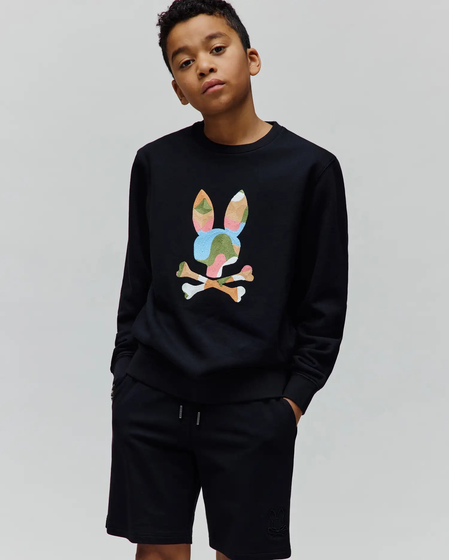 Black Kids Ara Crew sweatshirt featuring a large, vibrant multicolor camouflage Psycho Bunny skull logo on the chest, paired with black jersey shorts with a small tonal embroidered bunny logo on the left thigh, both in a relaxed fit.
