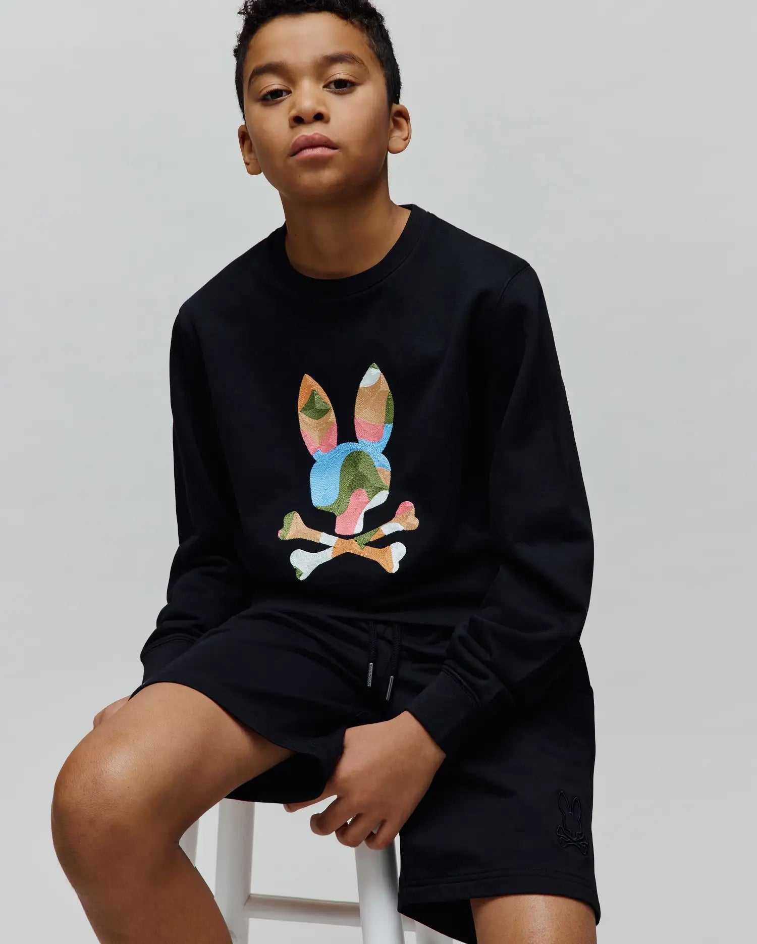 Black Kids Ara Crew relaxed fit french terry sweatshirt with a large multicolor camo embroidered Psycho Bunny skull logo on the chest, paired with black jersey shorts featuring a small tonal embroidered bunny skull on the left leg.