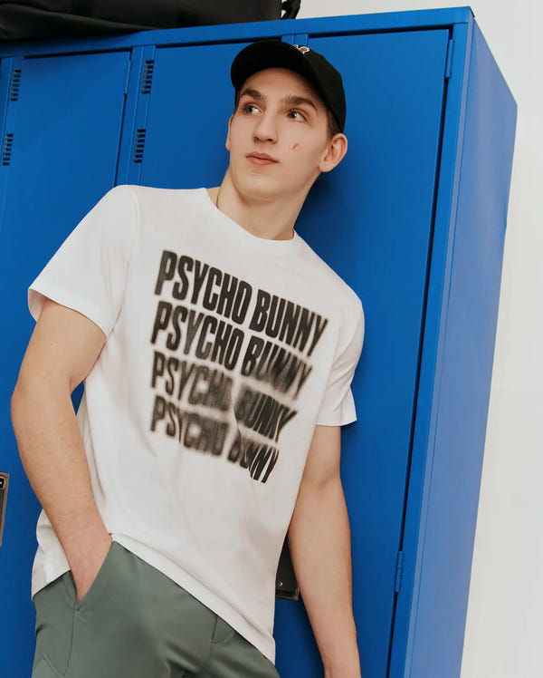 Mens Lawrence Graphic Tee, a white classic fit jersey t-shirt featuring a large, central black graphic of the word PSYCHO BUNNY repeated with a blurred, distorted effect.