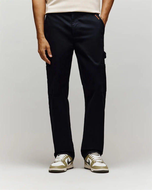 A person dressed in Psycho Bunny’s MENS RUSSELL CARPENTER PANT (MB0600005) with cargo pockets and white sneakers with olive green accents stands before a plain light background, visible only from the waist down.