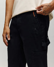 A person wears Psycho Bunny MENS RUSSELL CARPENTER PANT - MB0600005 in black, featuring an embroidered logo, orange stitching, and side cargo pockets; their tattooed left hand rests by their leg, paired with a light shirt.