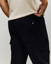 A person wearing a cream-colored shirt and black Psycho Bunny MENS RUSSELL CARPENTER PANT - MB0600005 stands with their back to the camera, one hand pulling at the waistband to show off these regular fit pants’ rear pocket details.