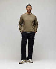 A man stands facing forward against a light background. He wears a khaki sweatshirt, Psycho Bunny MENS RUSSELL CARPENTER PANT - MB0600005 with cargo pockets, and beige sneakers with white accents. His hands are in his pockets and he looks neutral.