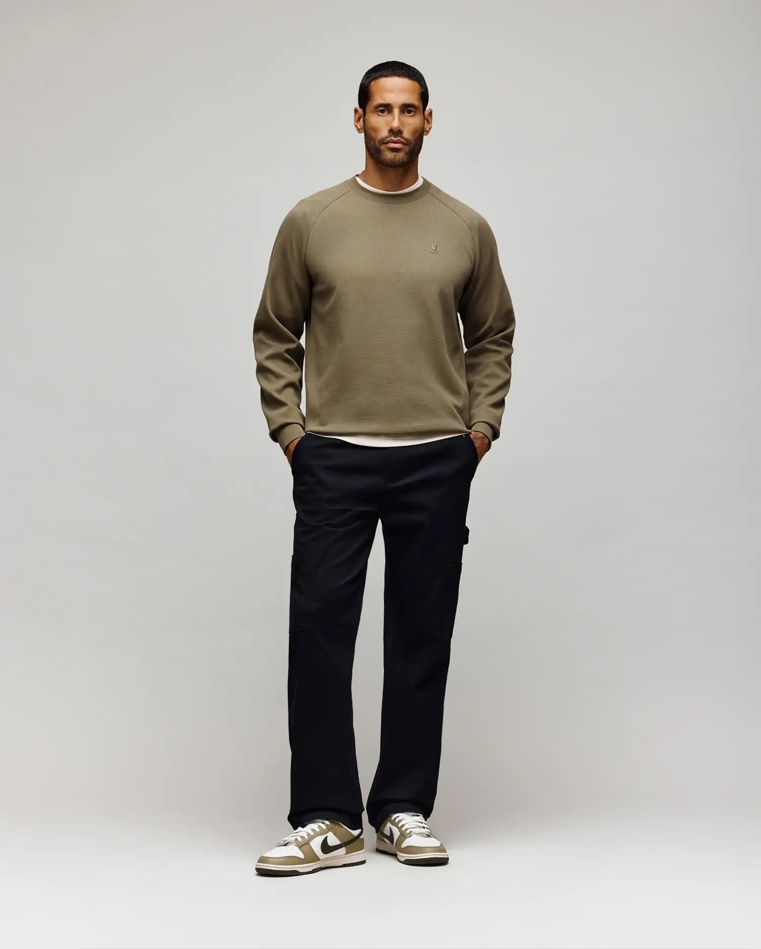 A man stands facing forward against a light background. He wears a khaki sweatshirt, Psycho Bunny MENS RUSSELL CARPENTER PANT - MB0600005 with cargo pockets, and beige sneakers with white accents. His hands are in his pockets and he looks neutral.