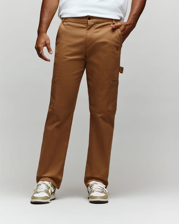 A person wearing Psycho Bunny MENS RUSSELL CARPENTER PANT - MB0600005 in brown, a white shirt, and white sneakers with gold accents stands against a plain light gray background. The upper body is mostly out of frame.