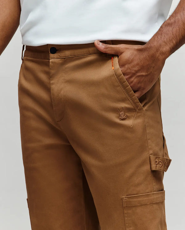 A person wears Psycho Bunny MENS RUSSELL CARPENTER PANT (MB0600005) in tan—regular fit, button closure, embroidered logo, visible "PSY" cargo pocket, and hammer loop—with one hand in their pocket and paired with a white shirt.