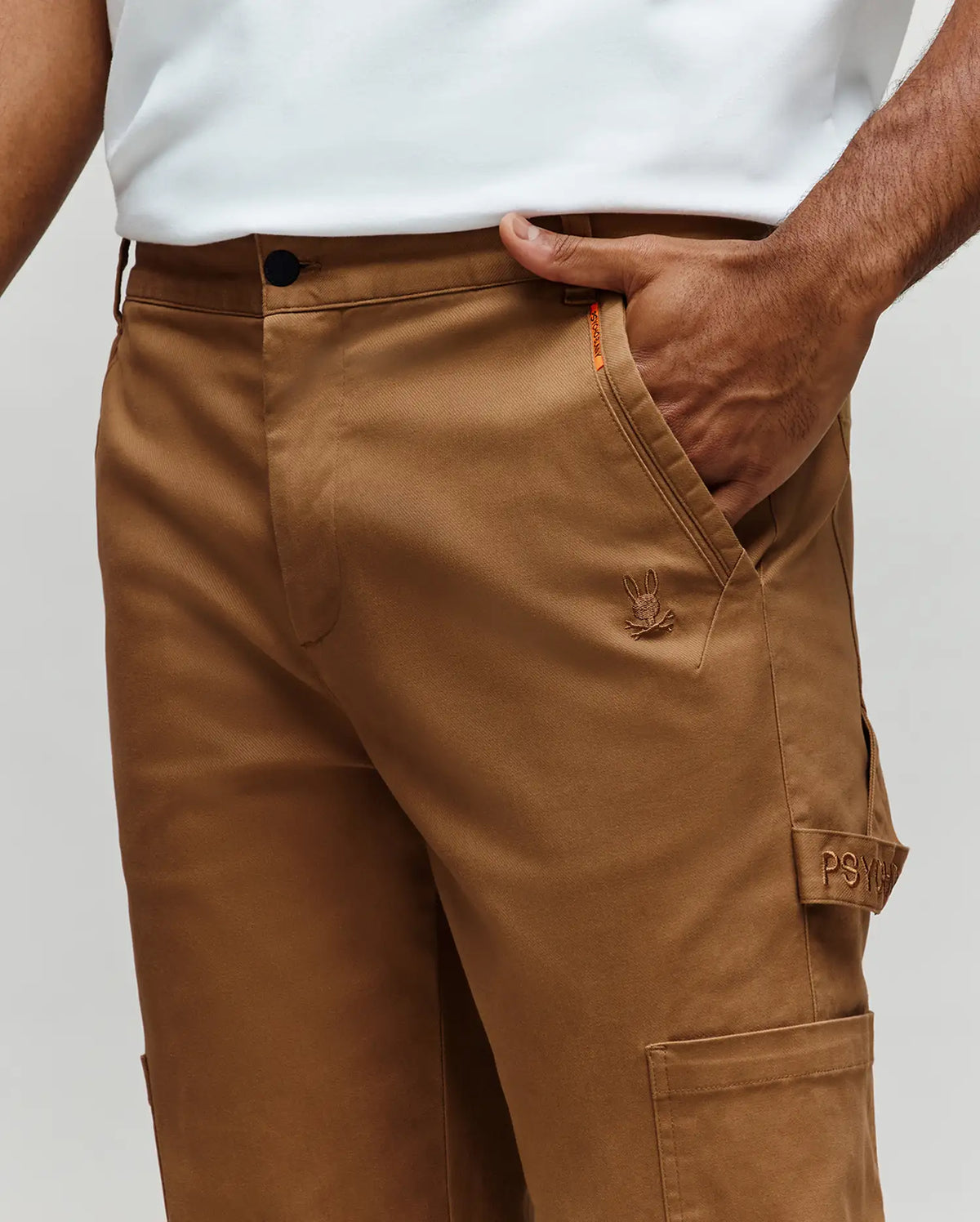 A person wears Psycho Bunny MENS RUSSELL CARPENTER PANT (MB0600005) in tan—regular fit, button closure, embroidered logo, visible "PSY" cargo pocket, and hammer loop—with one hand in their pocket and paired with a white shirt.