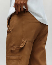 A person wears Psycho Bunny's MENS RUSSELL CARPENTER PANT - MB0600005 in brown, featuring back pockets, side details, hammer loop, and "PSYCHO" embroidery above a side flap. Their hand rests at the waistband over a crisp white shirt.