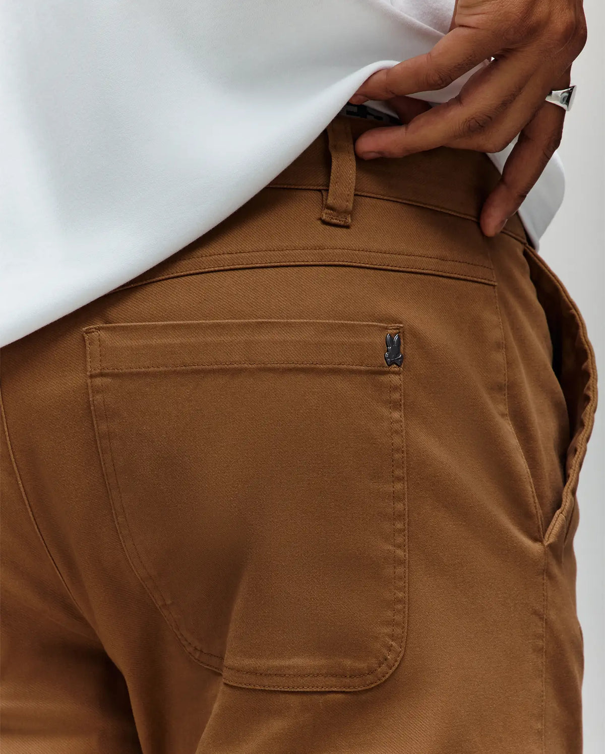 A close-up of a person wearing Psycho Bunny MENS RUSSELL CARPENTER PANT - MB0600005 in regular fit with a white shirt tucked in. Their hand, adorned with a silver ring, rests near the back pocket by the hammer loop.