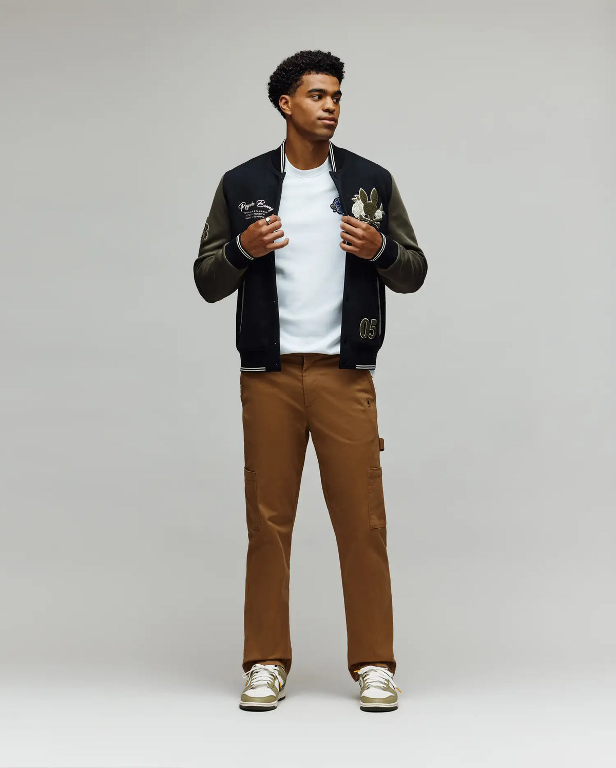 A man stands against a plain backdrop wearing Psycho Bunny's MENS RUSSELL CARPENTER PANT - MB0600005 with a hammer loop, styled with a black varsity jacket, white t-shirt, and white sneakers. He looks slightly aside, holding his jacket open.