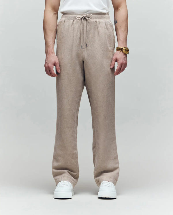 A person wears Psycho Bunny’s MENS FARRIS WIDE LEG LINEN PANT (MB0600007) in beige, styled with a white T-shirt, white sneakers, and a gold watch against a light gray background. The image is cropped from the shoulders down.