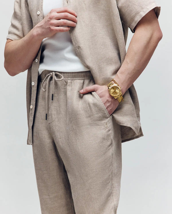 A person wears Psycho Bunny’s MENS FARRIS WIDE LEG LINEN PANT - MB0600007 in beige with a matching button-up shirt, white undershirt, and large gold watch. An embroidered logo is visible; their face is not shown.