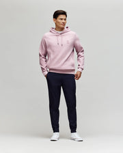 A man stands against a plain light background, wearing Psycho Bunny's MENS BEN TECH JOGGER PULL ON - MB0600013 in dark navy, paired with a light pink hoodie and white sneakers. He smiles slightly, hands in pockets, looking to the side.
