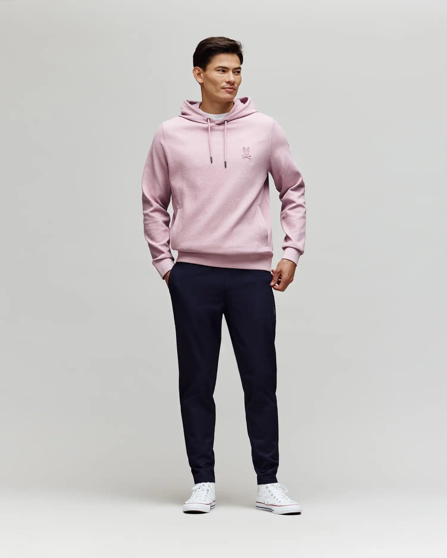 A man stands against a plain light background, wearing Psycho Bunny's MENS BEN TECH JOGGER PULL ON - MB0600013 in dark navy, paired with a light pink hoodie and white sneakers. He smiles slightly, hands in pockets, looking to the side.