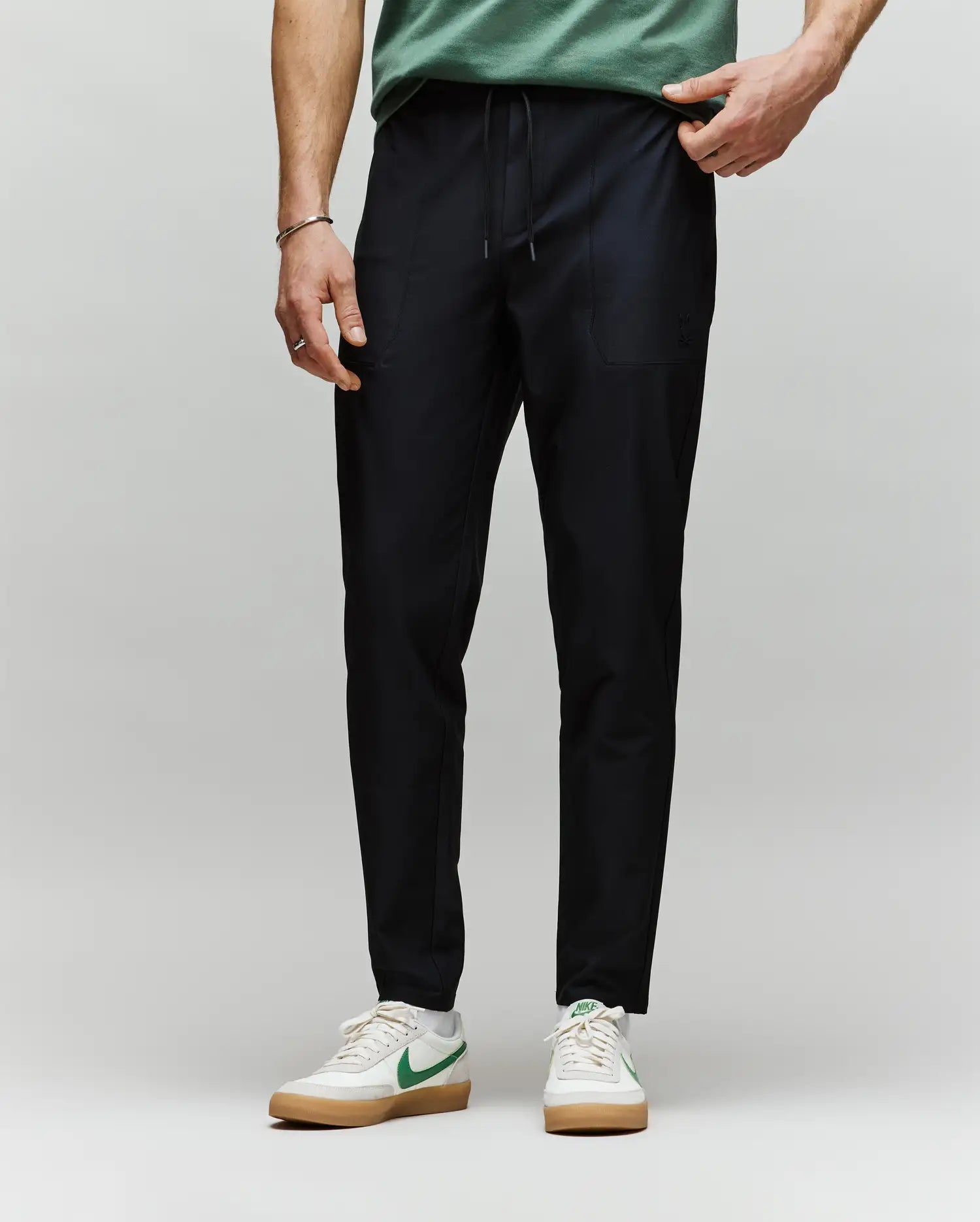 A person, visible from the waist down, stands against a plain background wearing Psycho Bunny MENS BEN TECH JOGGER PULL ON - MB0600013 in black, paired with a green shirt and white sneakers featuring green Nike swooshes.