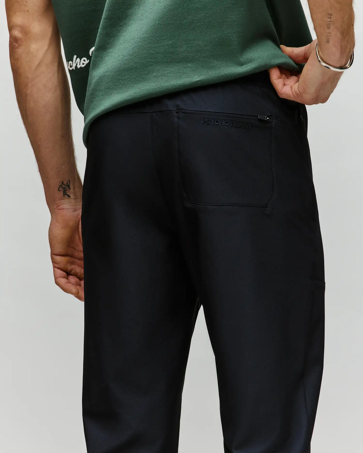 A person wearing a green t-shirt and dark "MENS BEN TECH JOGGER PULL ON - MB0600013" pants is shown from the back, one hand on their hip, with a back pocket featuring the Psycho Bunny brand name above it.