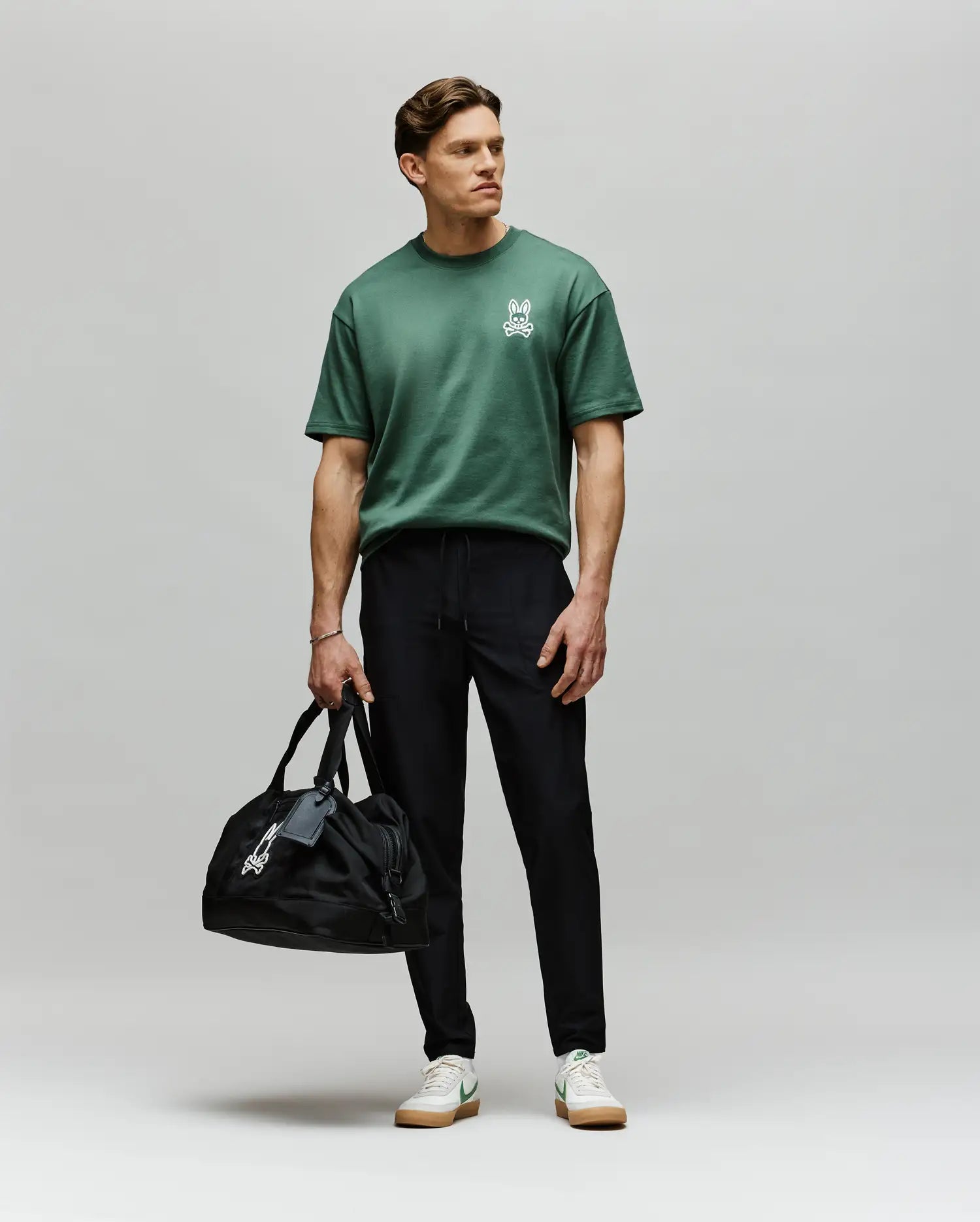 A man stands against a plain light background wearing Psycho Bunny MENS BEN TECH JOGGER PULL ON - MB0600013, a green polyester spandex t-shirt, and white sneakers. He holds a black duffel bag in his left hand and looks slightly to his right. Made in China.