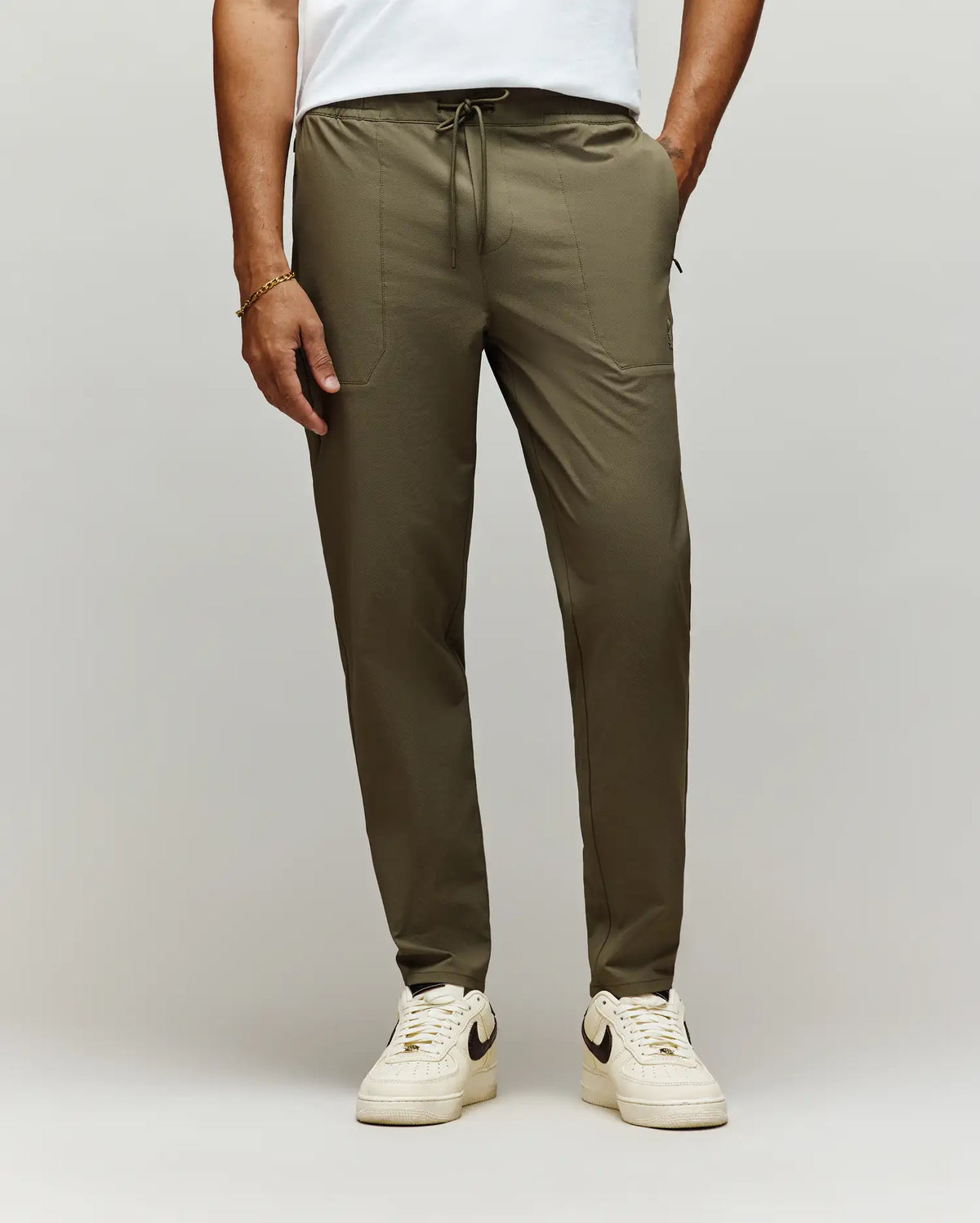 A man stands against a plain light background, showing only his lower body in Psycho Bunny MENS BEN TECH JOGGER PULL ON - MB0600013 (olive green), paired with a white t-shirt and white Nike sneakers.
