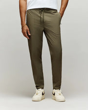 A man stands against a plain light background, showing only his lower body in Psycho Bunny MENS BEN TECH JOGGER PULL ON - MB0600013 (olive green), paired with a white t-shirt and white Nike sneakers.