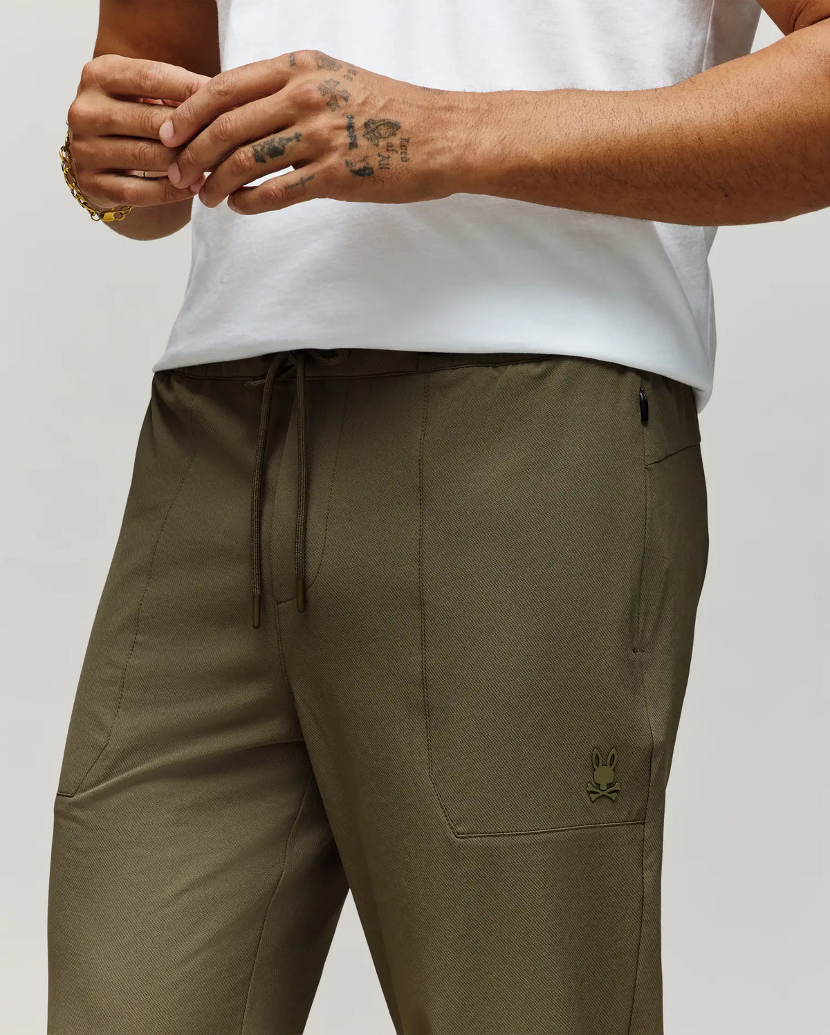 A person wears Psycho Bunny's MENS BEN TECH JOGGER PULL ON (MB0600013) in olive green, featuring a polyester-spandex blend, drawstring waist, and small bunny logo, paired with a white shirt; only torso/upper legs and one tattooed hand with bracelet are visible.