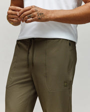 A person wears Psycho Bunny's MENS BEN TECH JOGGER PULL ON (MB0600013) in olive green, featuring a polyester-spandex blend, drawstring waist, and small bunny logo, paired with a white shirt; only torso/upper legs and one tattooed hand with bracelet are visible.