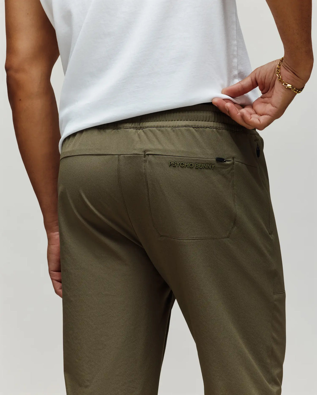 A man is shown from the back wearing Psycho Bunny's MENS BEN TECH JOGGER PULL ON (MB0600013) in olive green, featuring a rear pocket with "Psycho Bunny" embroidery, paired with a white shirt and a gold bracelet on his left wrist.