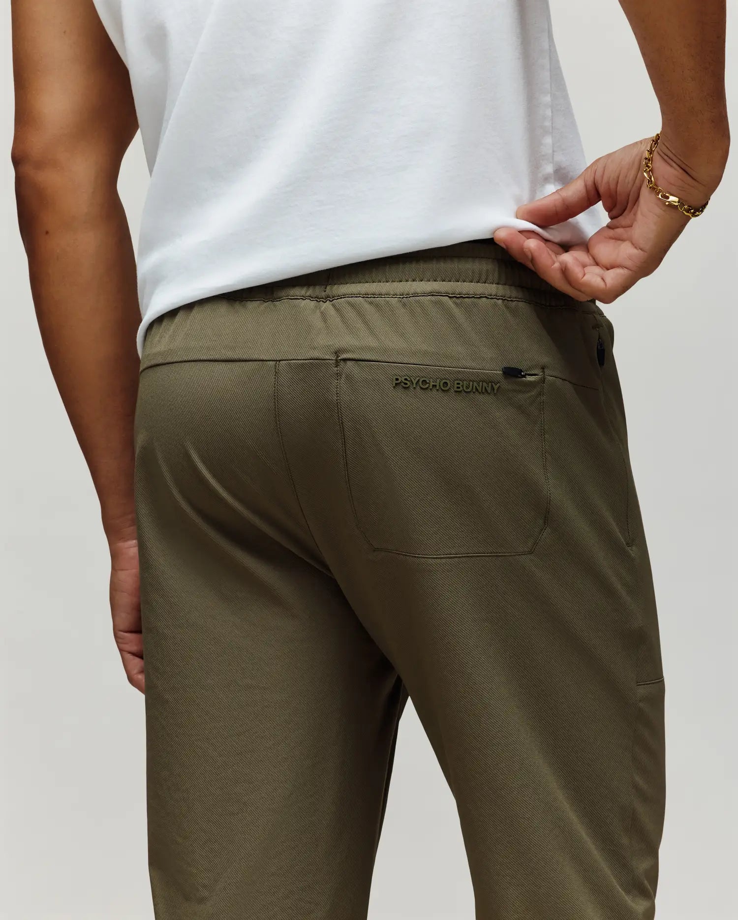 A man is shown from the back wearing Psycho Bunny's MENS BEN TECH JOGGER PULL ON (MB0600013) in olive green, featuring a rear pocket with "Psycho Bunny" embroidery, paired with a white shirt and a gold bracelet on his left wrist.