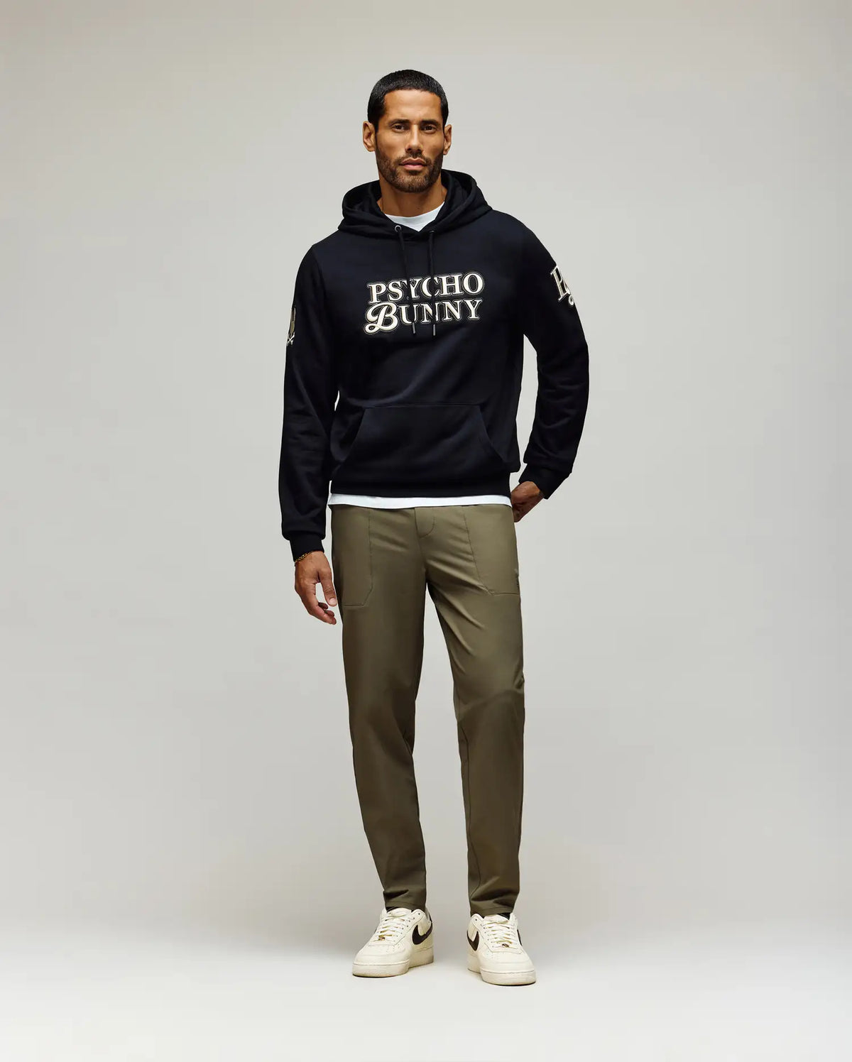 A man with short dark hair and a trimmed beard, looking slightly left, wears a black Psycho Bunny hoodie and olive green Psycho Bunny MENS BEN TECH JOGGER PULL ON - MB0600013 pants, paired with white sneakers.