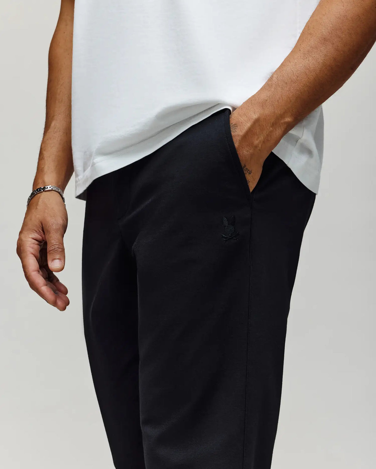 A person wears Psycho Bunny MENS EDWARD PANT - MB0600014, black cotton woven pants made in Vietnam, featuring a subtle embroidered logo on the hip. Only their lower torso and upper legs are visible, with one hand in their pocket.