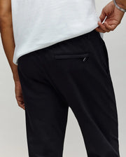 A person is shown from the waist down wearing Psycho Bunny MENS EDWARD PANT - MB0600014, black cotton woven pants, paired with a white shirt, with one hand holding the waistband near the back pocket.