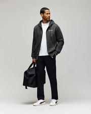 A man poses against a light gray background holding a black duffel bag, wearing Psycho Bunny's MENS EDWARD PANT - MB0600014 in black, paired with a dark gray zip-up hoodie, white t-shirt, and white sneakers.