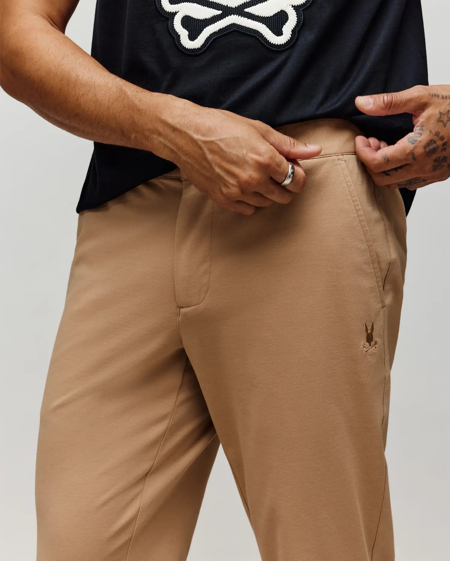 A person in Psycho Bunny MENS EDWARD PANT - MB0600014, tan cotton pants with a small embroidered logo on the pocket, adjusts the waistband to reveal a black shirt featuring a white skull and crossbones graphic.