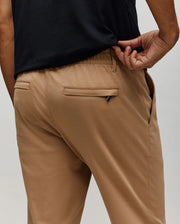 A person wearing Psycho Bunny MENS EDWARD PANT - MB0600014 in tan and a black shirt, shown from the back, lifts their shirt to reveal the waistband and zippered pocket on the cotton woven pants made in Vietnam.