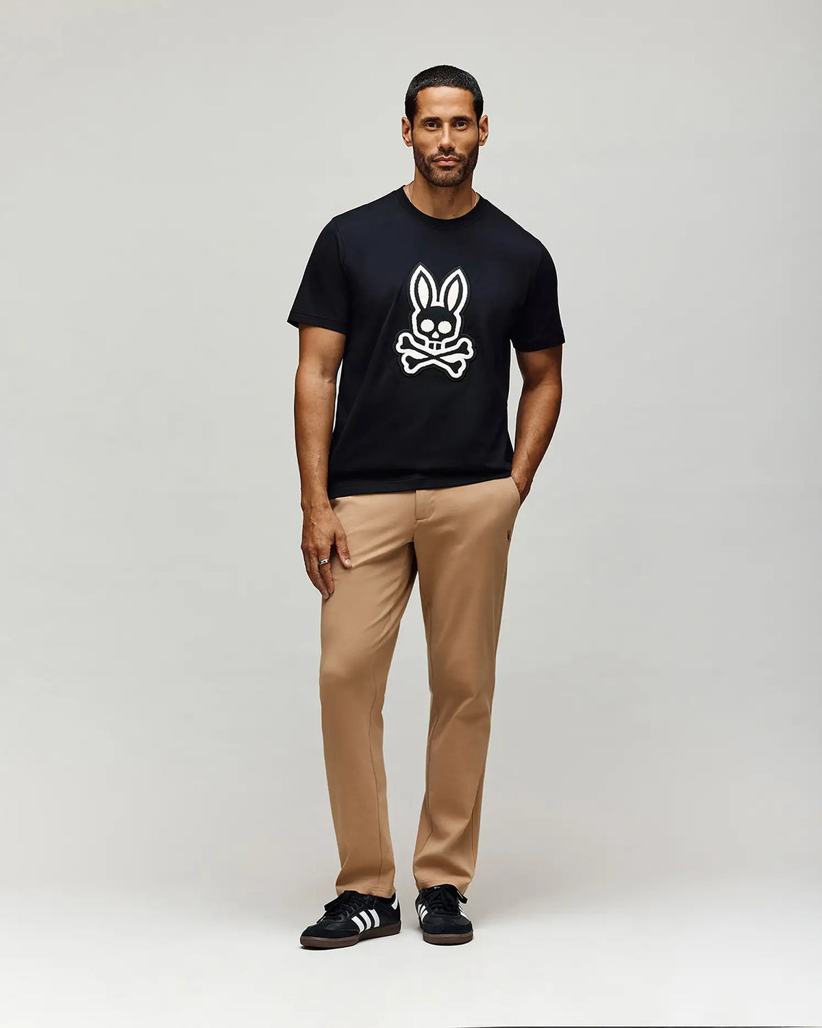 A man stands against a plain light background, wearing a black cotton woven t-shirt with a white bunny and crossbones graphic, MENS EDWARD PANT - MB0600014 by Psycho Bunny in tan, and black sneakers with white stripes.