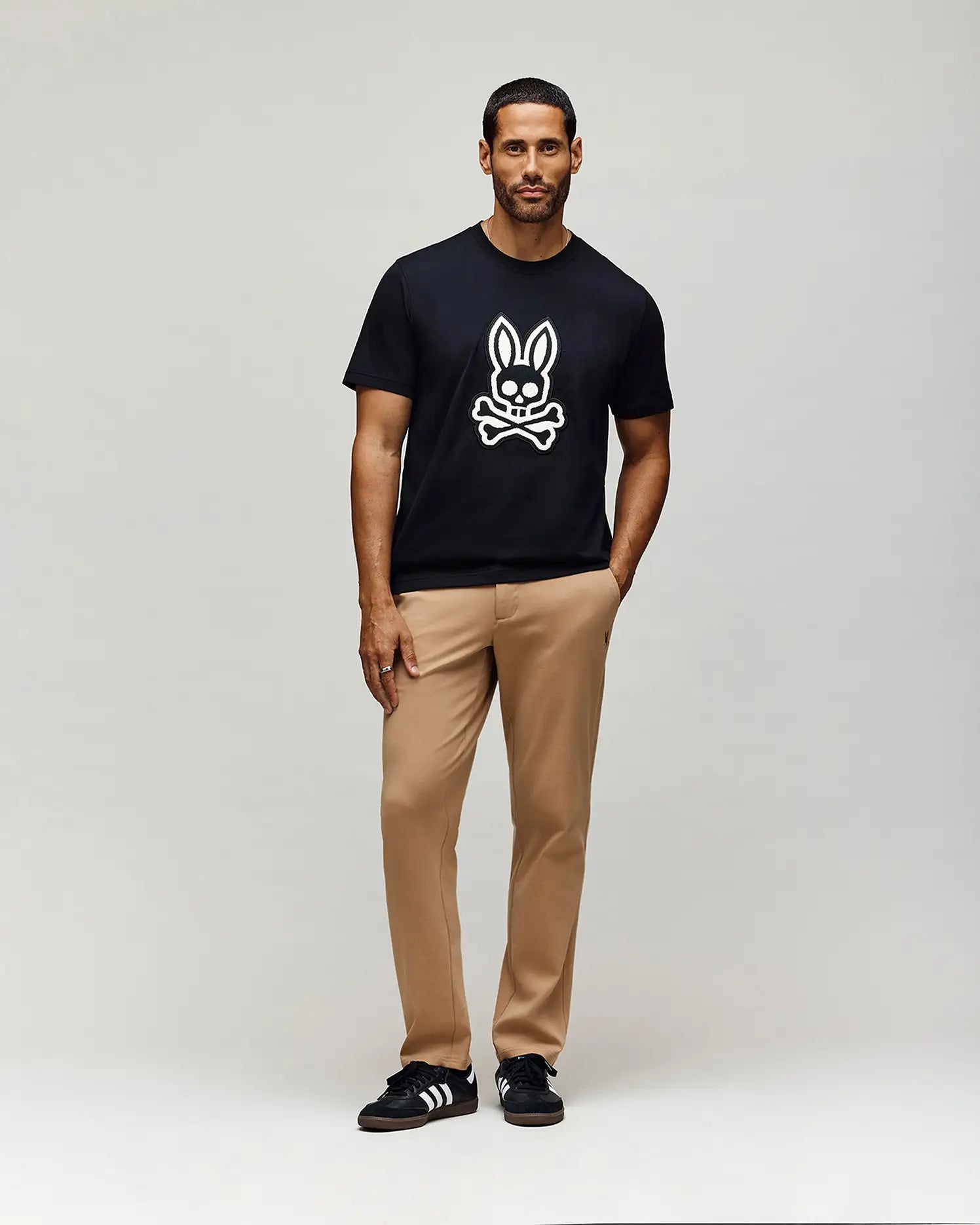 A man stands against a plain light background, wearing a black cotton woven t-shirt with a white bunny and crossbones graphic, MENS EDWARD PANT - MB0600014 by Psycho Bunny in tan, and black sneakers with white stripes.
