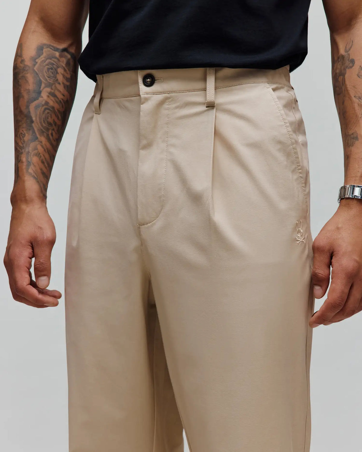 Mens Malik Wide Leg Pant in beige, featuring a relaxed fit, smooth woven fabric, single pleats, button closure, and a small embroidered Psycho Bunny skull logo near the right front pocket.