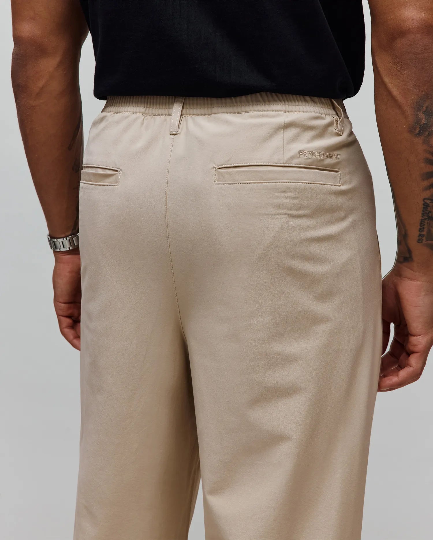 Mens Malik Wide Leg Pant in beige, featuring a relaxed fit, smooth fabric, elasticated waistband, two back welt pockets, and a small embroidered Psycho Bunny text logo above the right back pocket.