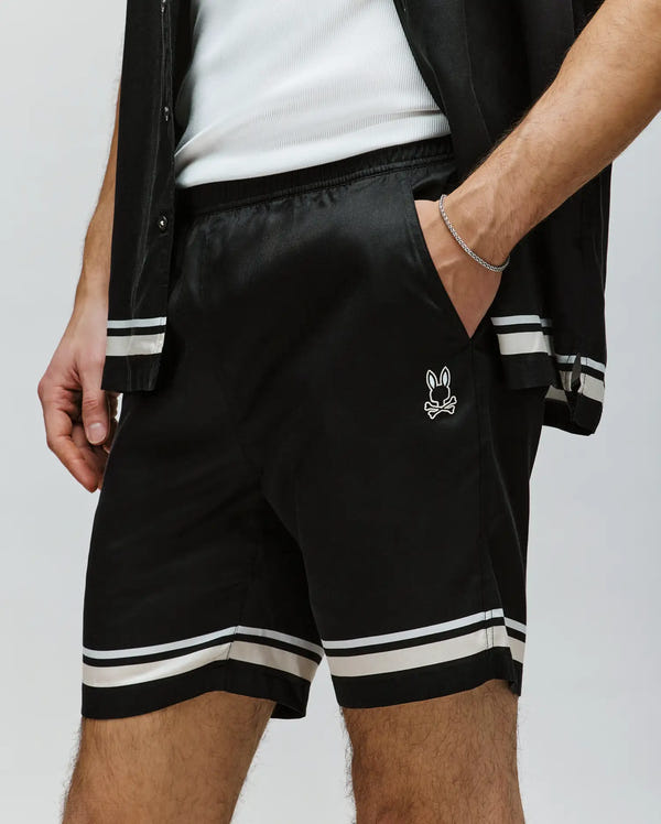 A person wears Psycho Bunny's MENS ADEN TENCEL SHORT (MB0700005) in black—100% tencel, with white stripes at the hem and a bunny skull logo—paired with a matching shirt and white tank, hand in pocket for a stylish, sustainable look.